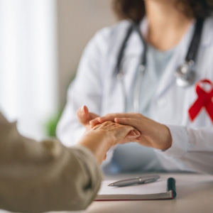 Close-up of a doctor holding a patient's hand with a red ribbon on the desk.