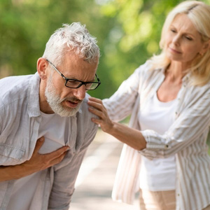 An elderly man clutching his chest while a woman provides support outdoors.