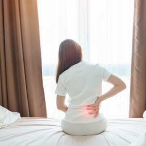 A woman sitting on a bed holding her lower back, illustrating lumbar spine pain.